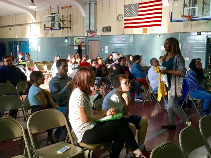 A group of adults and children in a school gym sit in chairs for a presentation.